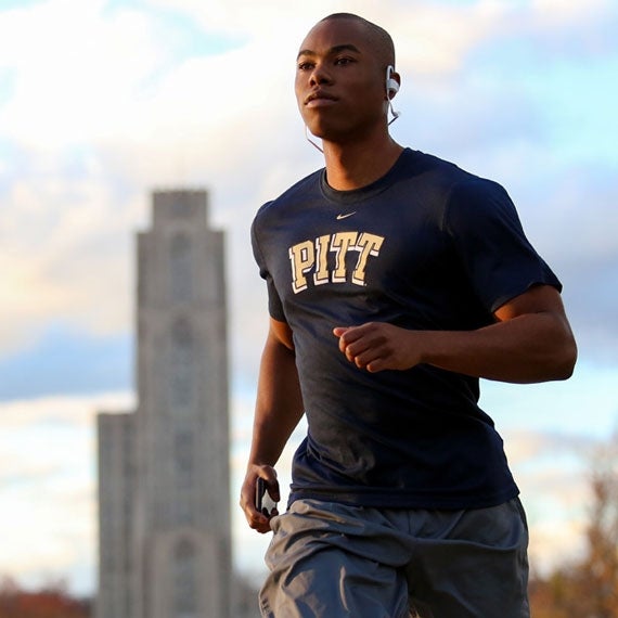 A young man wearing a Pitt shirt jogs with the Cathedral of Learning in the background